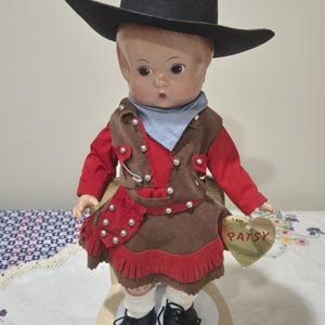 May include: A vintage Patsy doll wearing a black cowboy hat, red shirt, brown vest and skirt with red fringe. The doll has black shoes, white socks, and a blue neckerchief. A heart-shaped tag reads "I AM PATSY".