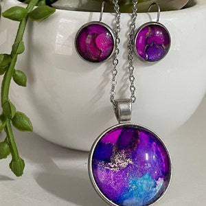 May include: A jewelry set featuring a pendant necklace and matching earrings. The pendant and earrings have a silver-tone metal frame and feature a vibrant abstract design in shades of purple, pink, blue, and gold.