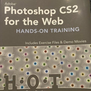 May include: A gray book with the title "Adobe Photoshop CS2 for the Web" and the subtitle "Hands-On Training". The book cover features a pattern of colorful eyes and the text "HOT" in large black letters. The author's name, "Tanya Staples", is printed below the text.