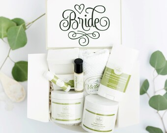 May include: A white gift box with a card that says "Bride" on it. The box is filled with various bath and body products, including a white towel, a white jar of body cream, a white jar of hand cream, a white deodorant, a white lip balm, and a small bottle of essential oil.