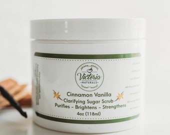 May include: A white jar of Cinnamon Vanilla Clarifying Sugar Scrub by Victoria Naturals. The jar is labeled with the product name and description: "Clarifying Sugar Scrub, Purifies - Brightens - Strengthens, 4oz (118ml)".  A vanilla bean and cinnamon sticks are in the foreground.
