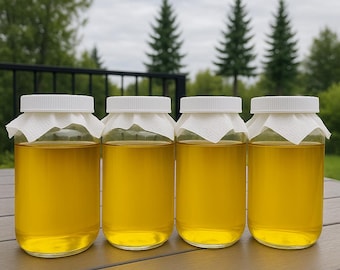 May include: Four clear glass jars filled with a golden-yellow liquid, each sealed with a white lid and a paper covering. The jars are arranged on a brown wooden surface, with a blurred outdoor background of trees and a cloudy sky.