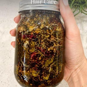May include: A clear glass jar filled with Ayurvedic hair oil, containing various herbs and botanicals. The jar has a silver lid and is held by a hand. Text on the image reads "Ayurvedic Hair Oil | Natural Growth & Strong Hair Care".