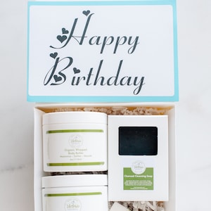May include: A white gift box with a blue lid. The lid has a card that says "Happy Birthday" with hearts. The box contains four bath and body products: a jar of organic whipped body butter, a jar of coffee oat scrub, a bar of charcoal cleansing soap, and a tube of lip balm. All products have the same brand name, "Victoria Naturals."