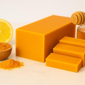 May include: An arrangement of natural ingredients for skincare. Includes a large block of orange soap, three smaller bars, a wooden bowl of turmeric powder, a lemon slice, and a jar of honey with a honey dipper.