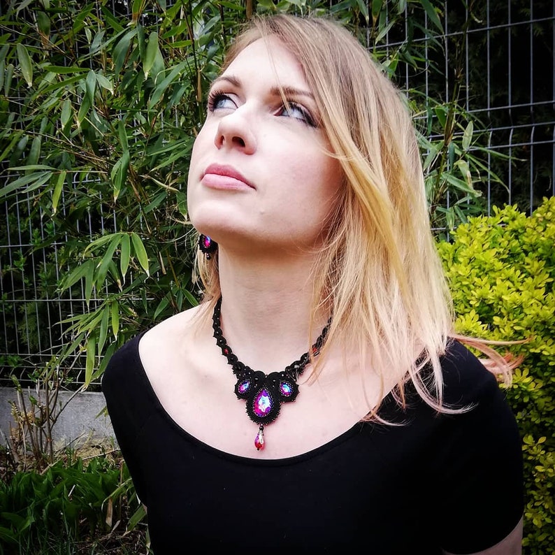 May include: A woman wearing a black top with a black beaded necklace featuring pink and blue iridescent stones. The necklace has a teardrop pendant.