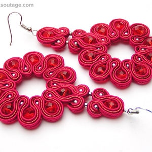 May include: A pair of pink earrings with red crystal accents. The earrings are made of a woven material and have a floral design.