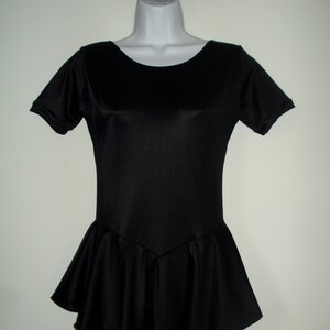 May include: A black leotard with short sleeves and a flared skirt, displayed on a mannequin. A matching black scrunchie is attached to the hanger. The leotard has a scoop neckline and a fitted bodice.