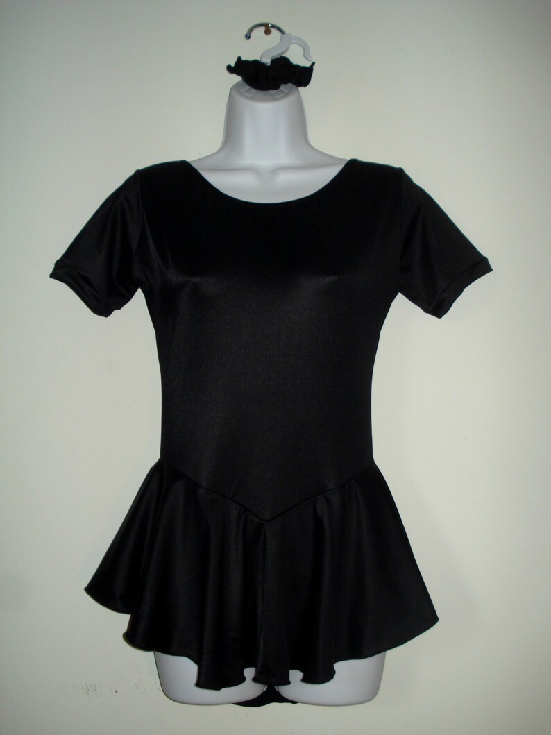 May include: A black leotard with short sleeves and a flared skirt, displayed on a mannequin. A matching black scrunchie is attached to the hanger. The leotard has a scoop neckline and a fitted bodice.