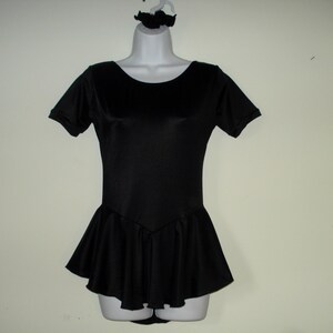 May include: A black leotard with short sleeves and a flared skirt, displayed on a mannequin. A matching black scrunchie is on the hanger above the leotard. The leotard has a scoop neckline and a fitted bodice.