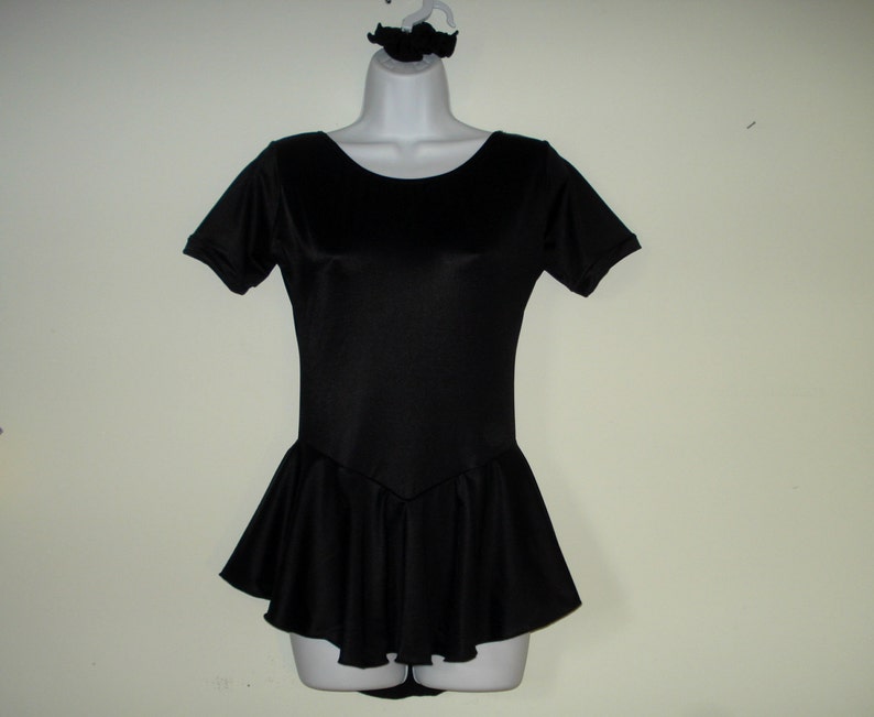 May include: A black leotard with short sleeves and a flared skirt, displayed on a mannequin. A matching black scrunchie is on the hanger above the leotard. The leotard has a scoop neckline and a fitted bodice.