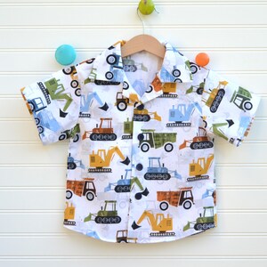 May include: White short-sleeved button-down shirt with a pattern of colorful construction vehicles. The shirt has a collar and a button closure.