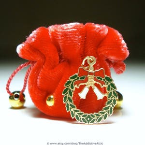 May include: A small, red velvet pouch with a drawstring closure and gold-coloured accents. A gold-toned Christmas wreath charm with green leaves, a red bow, and white bells hangs from the pouch. The pouch has two gold-coloured bells.