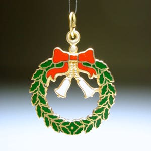 May include: A gold-coloured Christmas wreath pendant featuring a red bow and white bells. The wreath is green with gold trim. The pendant is suspended by a thin gold chain, ideal for festive jewellery.