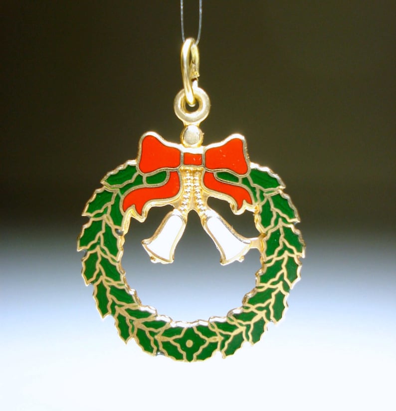 May include: A gold-coloured Christmas wreath pendant featuring a red bow and white bells. The wreath is green with gold trim. The pendant is suspended by a thin gold chain, ideal for festive jewellery.