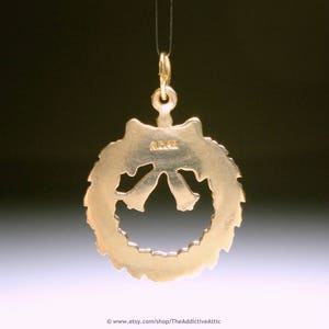 May include: A gold-coloured pendant shaped like a wreath with a bow at the bottom, suspended from a thin gold chain. The text "A.G. 14K" is visible on the pendant. The background is a gradient of dark and light colours.