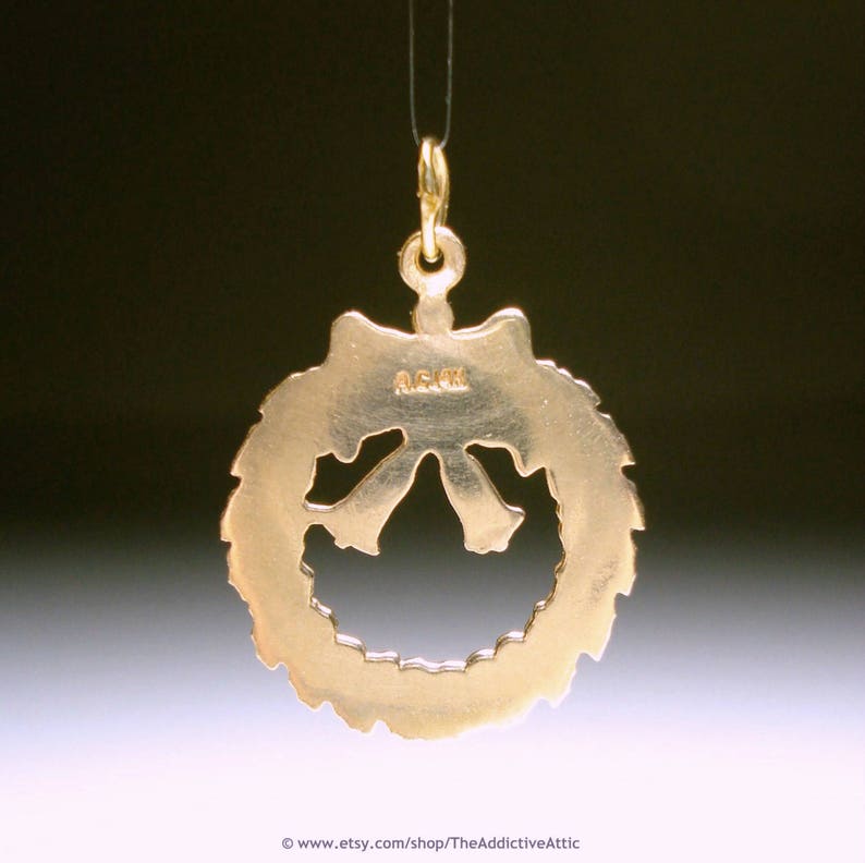 May include: A gold-coloured pendant shaped like a wreath with a bow at the bottom, suspended from a thin gold chain. The text "A.G. 14K" is visible on the pendant. The background is a gradient of dark and light colours.