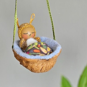 Puede incluir: Una pequeña muñeca con un gorro de punto amarillo y marrón y una manta con estampado floral está durmiendo en una hamaca de cáscara de nuez.