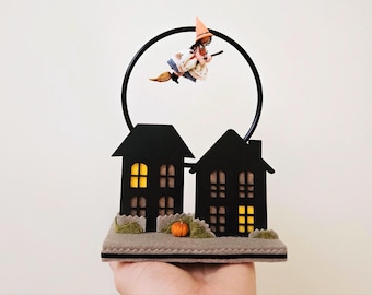 May include: A black and white Halloween-themed decoration with two houses, a witch flying on a broomstick, and a pumpkin. The houses have windows with yellow lights inside.