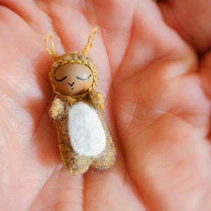 May include: A small, handmade doll with a brown wooden head and closed eyes. The doll is wearing a yellow and brown felt outfit with white felt accents. The doll is lying on its back in the palm of a hand.
