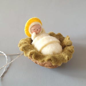 May include: A felt baby Jesus ornament in a manger made of a walnut shell. The baby is wearing a yellow halo and a white robe. The manger is surrounded by green felt.
