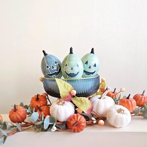 Puede incluir: Tres calabazas verdes con caras de calabaza tallada se muestran en un tazón azul con pedestal. Las calabazas están rodeadas de una guirnalda de hojas verdes, calabazas blancas y calabazas naranjas.