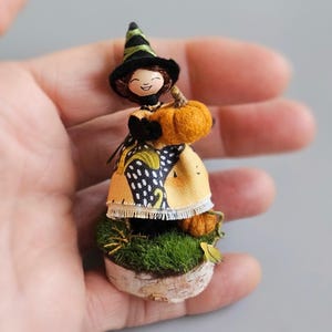 May include: A small witch doll wearing a black and green hat, a yellow and black polka dot dress, and holding a small orange pumpkin. The doll is standing on a small wooden base covered in green moss.