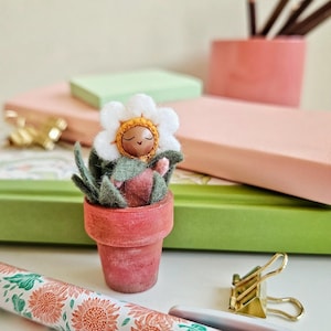 Puede incluir: Una muñeca de fieltro con forma de flor en una maceta de terracota, con una cabeza de flor blanca en forma de margarita y hojas verdes. La muñeca está colocada sobre un escritorio con cuadernos, un rollo de papel con estampado floral y un clip dorado.