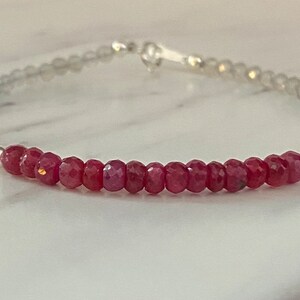 May include: A delicate bracelet featuring a central row of small, faceted, deep red beads flanked by two silver-toned spacer beads. The remainder of the bracelet consists of translucent, light grey, faceted beads, finished with a silver-toned clasp.
