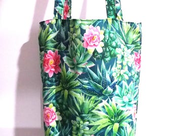May include: A tote bag with a botanical print featuring green succulents and pink flowers. The bag has a dark teal background and matching handles. The bag is hanging against a white background.