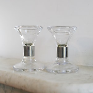 May include: Two clear glass candle holders with silver bands around the base. The candle holders are on a white marble surface.