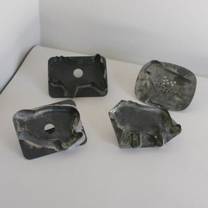 May include: Four vintage metal cookie cutters shaped like animals. The cutters are made of a dark gray metal and have a worn, antique look. The shapes include a pig, a cow, a lion, and a sheep.