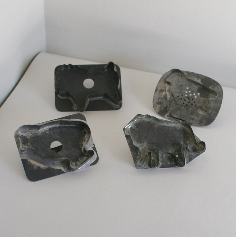 May include: Four vintage metal cookie cutters shaped like animals. The cutters are made of a dark gray metal and have a worn, antique look. The shapes include a pig, a cow, a lion, and a sheep.