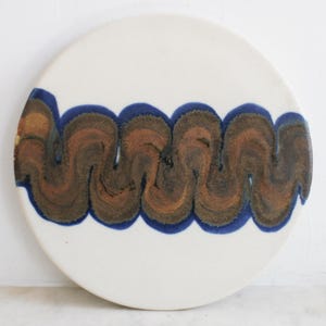 May include: A round, white ceramic plate with a wavy, abstract design. The design features a brown and blue pattern across the center of the plate. The plate has a glossy finish and is suitable for serving food.