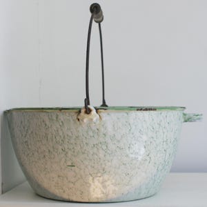 May include: An antique enamelware pot with a speckled white and green pattern. It features a metal handle with a wooden grip and a green rim. The pot has a rounded shape and a small handle on the side.