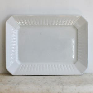 May include: A rectangular white ceramic serving platter with a slightly raised rim and a ribbed design on the edges. The platter has a clean, minimalist aesthetic, suitable for serving food. The dimensions are approximately 12 inches long and 8 inches wide.