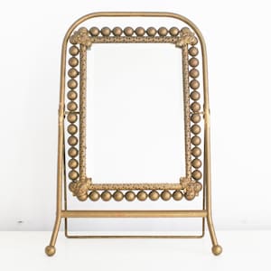 May include: A gold-colored vanity mirror with a rectangular frame. The frame is adorned with small, round embellishments and decorative floral accents. The mirror is supported by a stand with a curved top and a horizontal bar.