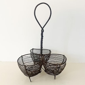 May include: A dark brown metal three-tiered basket with a handle. The basket has three round compartments supported by thin legs. The handle is a twisted design that ends in a loop.