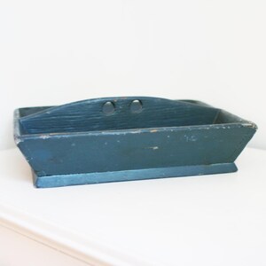 May include: A vintage, rectangular wooden tray painted in a distressed teal blue. The tray features a curved handle with two circular cutouts and a raised base. The paint shows signs of wear, adding to its rustic charm.