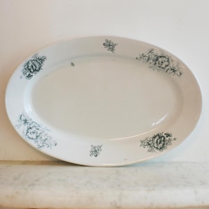 May include: Oval white serving platter with a floral design in teal. The platter has a smooth surface and a slightly raised edge. The floral pattern is repeated around the rim of the platter. A vintage piece for serving food.
