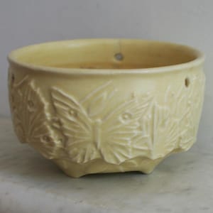 May include: A light yellow ceramic planter with a butterfly and leaf relief design. The planter has a round shape with a slightly flared rim and a series of small holes near the top. The planter is designed to hold plants.