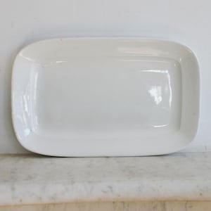 May include: A rectangular, white ceramic serving platter with rounded corners. The platter has a glossy finish and is empty. The background is a white wall and a marble surface.