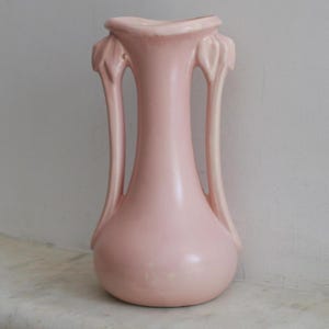 May include: A pale pink ceramic vase with a bulbous base and a slender neck. The vase features two curved handles and decorative floral accents near the top. The vase is approximately 10 inches tall.