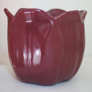 May include: A burgundy ceramic tulip-shaped planter. The planter has a glossy finish and a scalloped rim. The sides of the planter curve inward, creating a tulip-like shape. The planter is designed to hold plants or flowers.