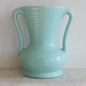 May include: A light blue ceramic vase with two curved handles. The vase has a wide base and a flared top, with horizontal ridged detailing around the body. The handles curve gracefully from the top to the body of the vase.