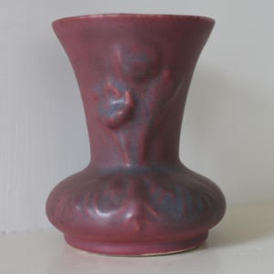 May include: A maroon ceramic vase with a bulbous base and flared top. The vase features embossed floral designs and a textured surface. The color is a deep reddish-purple with subtle blue accents, creating a vintage aesthetic. The vase is approximately 8 inches tall.