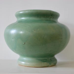 May include: A small, rounded ceramic vase with a smooth, light green glaze. The vessel features a wide, bulbous body that tapers toward a flared base and a short, thick neck with a rounded rim. The surface shows subtle variations in color.