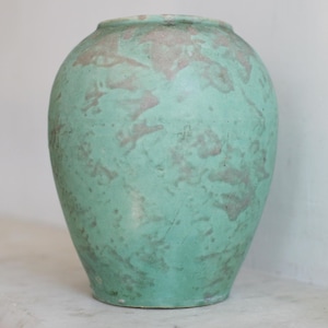 May include: A large, round, turquoise-colored ceramic vase with a textured surface. The vase has a wide body that tapers towards the top and bottom. The surface has a mottled appearance with patches of a lighter, grayish-pink color.