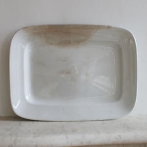May include: A rectangular white ceramic serving platter with rounded corners. The platter has a slightly raised edge and a brown stain on one side. The surface is smooth and glossy, reflecting the light.