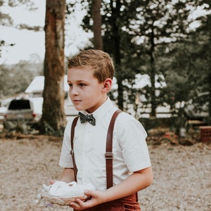 May include: A young boy wearing a white button-down shirt with brown braces and a grey bow tie holds a white cushion in his hands. He is standing on a gravel path in front of a wooded area.